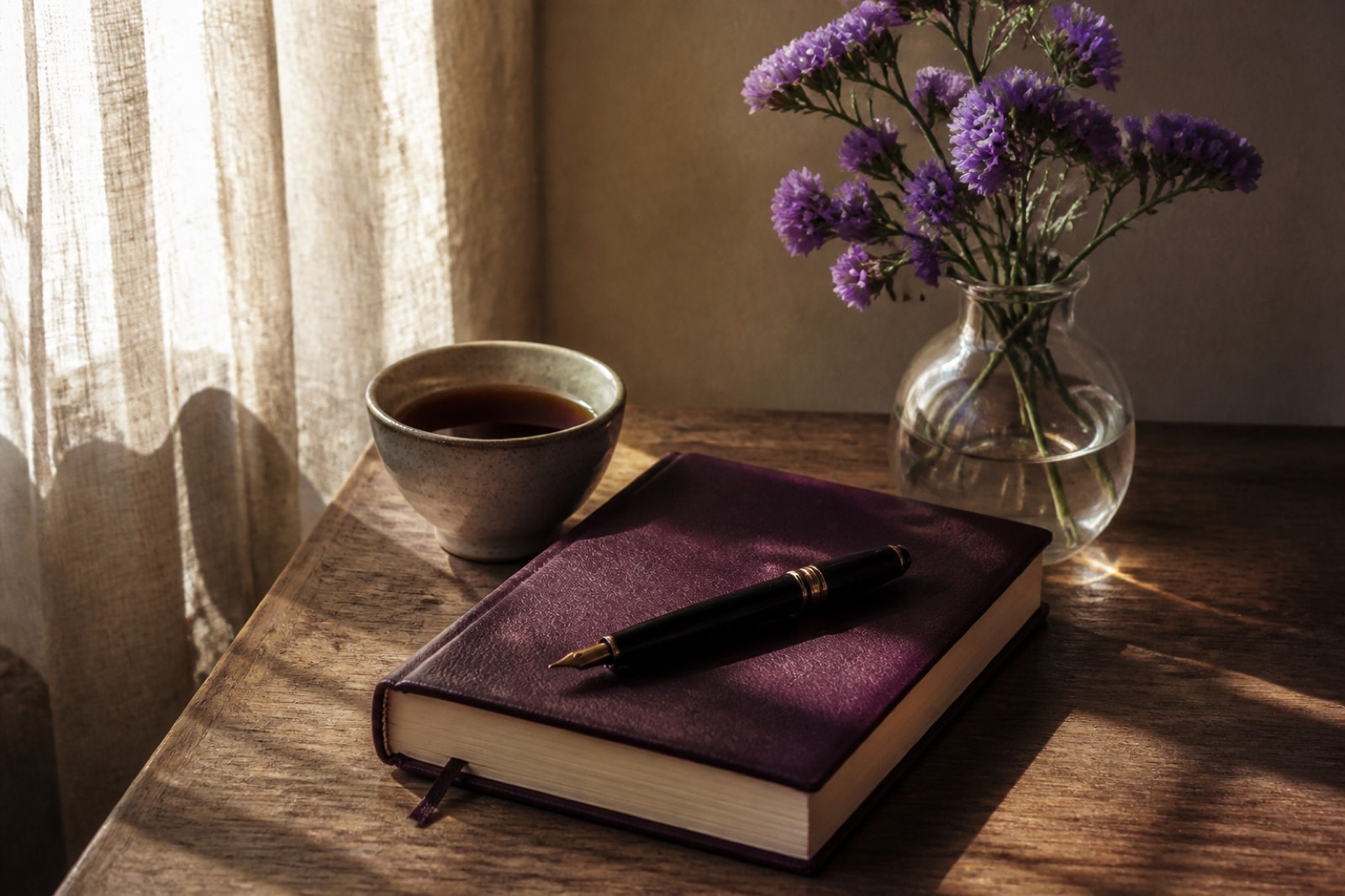 A morning desk with a plum notebook, fountain pen and a small vase of purple statice
